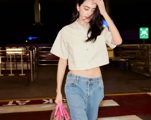  Mai Davika lăng xê quần jeans cạp trễ, áo sơ mi crop top và sneakers khi di chuyển ra sân bay cùng chiếc túi oversized màu hồng họa tiết monogram. 